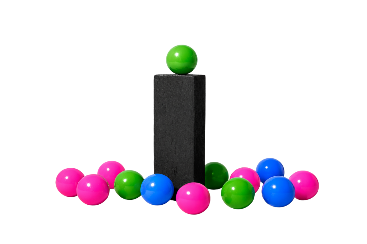 Minimal black and white still life showing a tall rectangular block at the center with a single green sphere balanced on top, surrounded by scattered spheres in bright pink, green, and blue on a white surface against a clear background.